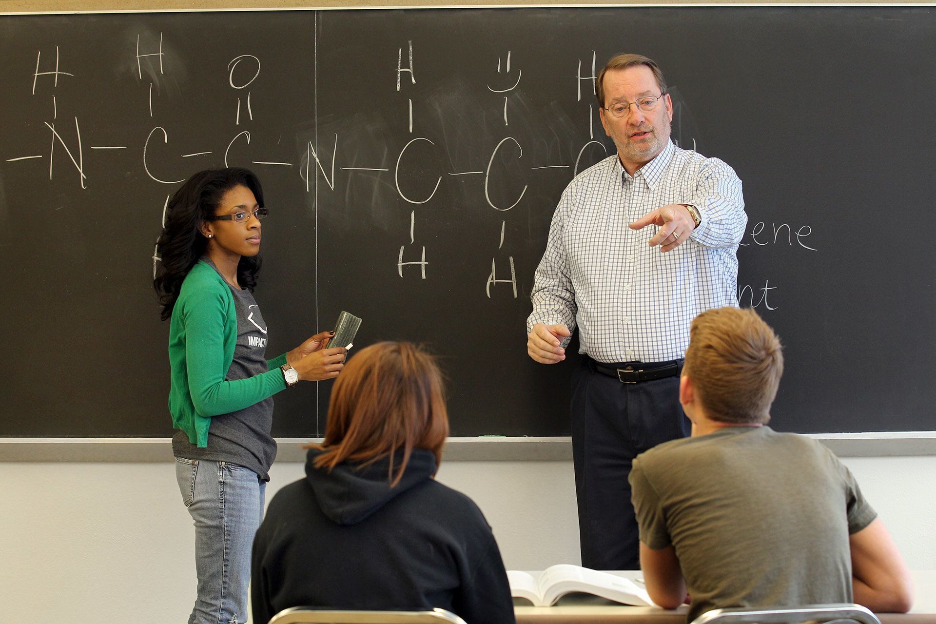 Two people stand in front of a chalkboard and two students. Two people stand in front of a chalkboard and two students.